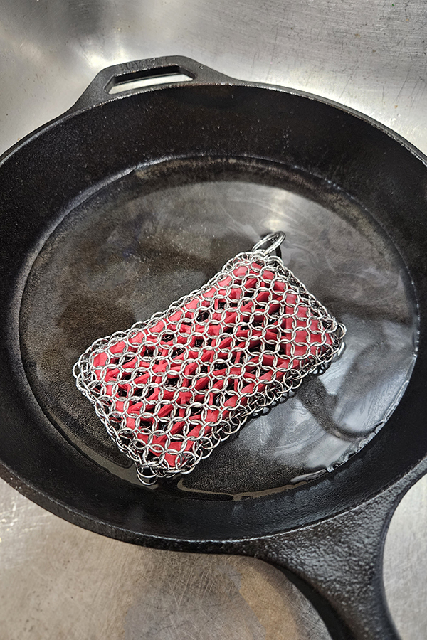 Water in a cast iron skillet and a red  chainmail scrubber in a sink.
